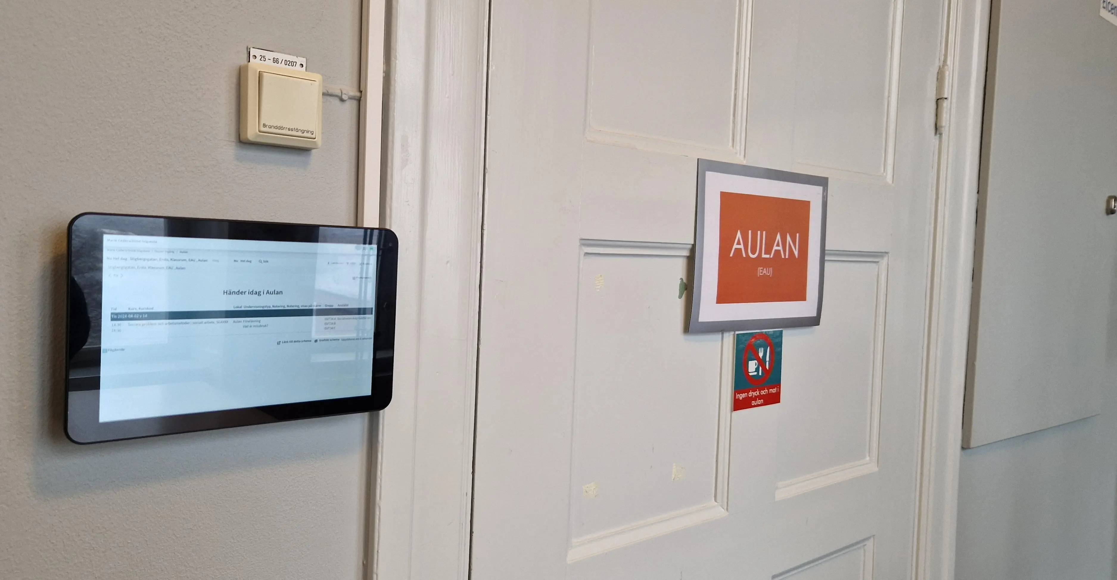 En svart skärm sitter placerad utanför Aulan. Bilden visar skärmen och Aulans dörr med en orange skylt där salsnamnet står.